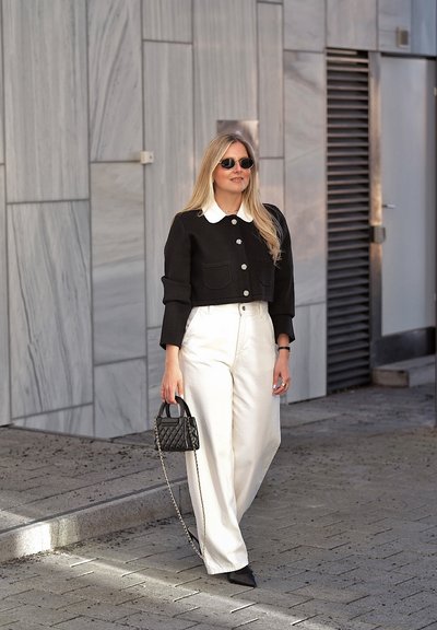 Mujer con chaqueta negra corta, pantalones blancos de cintura alta, zapatos negros puntiagudos, gafas de sol y sosteniendo un pequeño bolso negro acolchado caminando al aire libre.