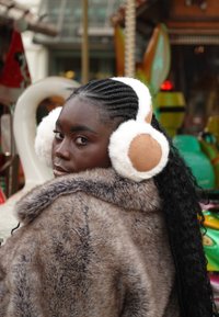 Manteau en fausse fourrure avec un motif texturé gris ; le modèle porte des cache-oreilles duveteux blancs et marron clair, mettant en valeur une coiffure élégante en tresse sur un fond coloré.