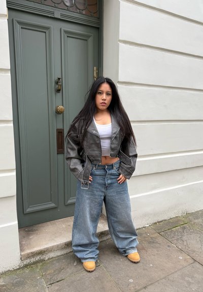 Chaqueta de cuero negra corta, top corto gris, jeans de mezclilla azules holgados y calzado color tostado. De pie frente a una puerta verde con vidrio decorativo.