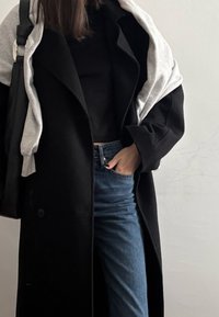 Femme portant un manteau noir par-dessus un haut noir et un jean bleu, avec un sweat-shirt gris drapé sur les épaules et un sac noir sur un côté.