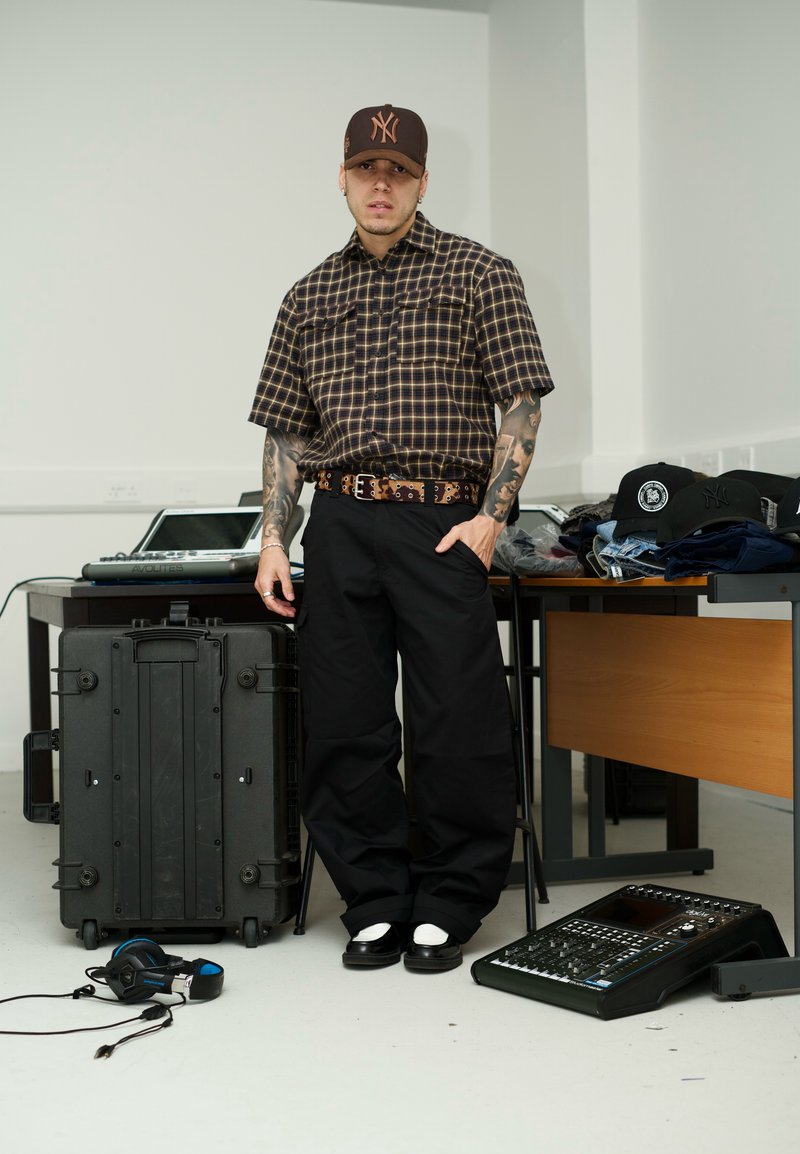 A person wearing a brown checked short-sleeve shirt, black wide trousers, and white trainers, standing next to a black case and a music device.
