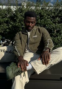 Ελιόχρωμο φερμουάρ μπουφάν με λευκές λεπτομέρειες, μπεζ παντελόνι και σκούρα πράσινα αθλητικά παπούτσια. Καθισμένος σε φόντο με πράσινη βλάστηση.