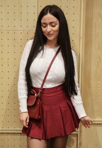 Biely sveter s kabelovým vzorom, bordová plisovaná sukňa a malá červená kabelka cez rameno. Hladké textúry a jasný farebný kontrast na drevenom pozadí.