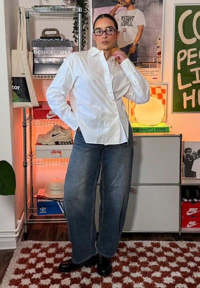 Mujer con gafas, camisa blanca abotonada, vaqueros anchos, zapatos negros, de pie en una habitación con zapatillas, carteles y una alfombra con estampado.