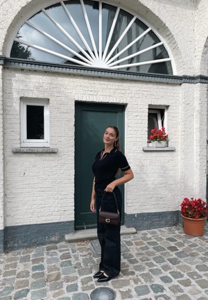 Polo negro con detalles en blanco, jeans de pierna ancha oscuros y un bolso marrón. Situado frente a una pared blanca texturizada con una puerta verde y flores rojas.