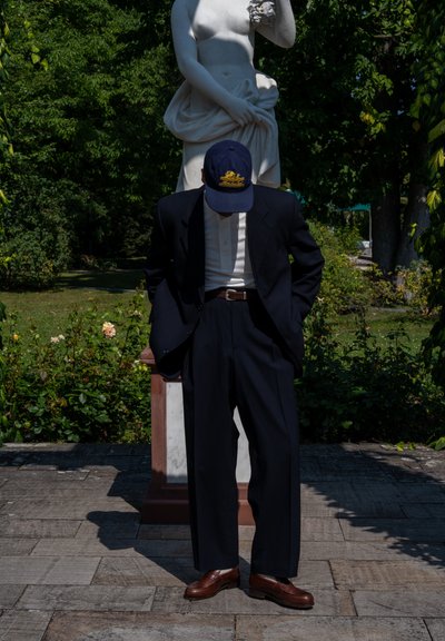 Traje de marino con pantalones de pierna ancha, camisa blanca debajo, zapatos de cuero marrón y una gorra azul con un emblema dorado, con fondo de estatua.