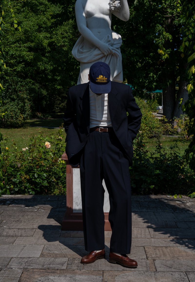 Completo blu con pantaloni a gamba larga, camicia bianca sotto, scarpe in pelle marrone e un cappello blu con un emblema dorato, il tutto davanti a uno sfondo di statua.