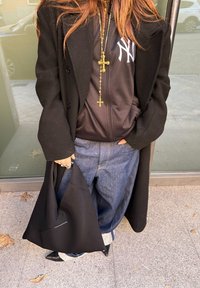 Μαύρο παλτό, μαύρη φούτερ με λευκό λογότυπο, χρυσό σταυρό κρεμαστό, μπλε τζιν, και μαύρη γωνιακή τσάντα. Ορατά μαύρα τακούνια και γκρι κάλτσες.