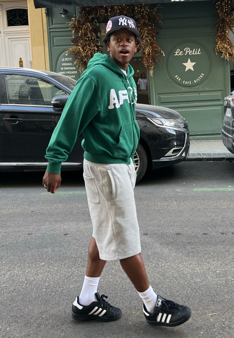 Sweat-shirt vert avec des lettres blanches, shorts gris, chaussettes blanches et baskets noires avec des rayures blanches. Scène sur le trottoir avec une voiture en arrière-plan.