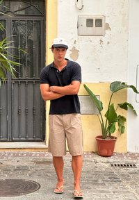 Cămașă polo neagră, pantaloni scurți bej și șlapi albi. Stând în fața unui perete texturat, cu vegetație și o poartă metalică în spate.