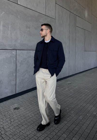 Chaqueta de cremallera navy sobre un polo oscuro, pantalones de pierna ancha beige y zapatos negros. Tejido suave y diseño estructurado con dos bolsillos en el pecho.