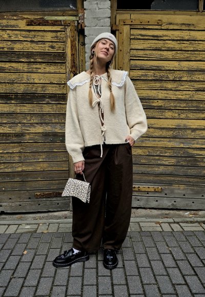 Cárdigan de punto en color crema con cuello de encaje, atado en la parte delantera; combinado con pantalones de pierna ancha de rayas finas en color oscuro. Pequeño bolso de mano con estampado de leopardo. Zapatos negros.