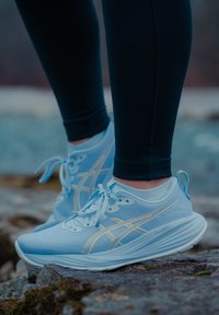 Pieds chaussés de baskets de course bleu clair à semelles blanches, posés sur un sol rocheux, portant des leggings noirs et des chaussettes blanches aux chevilles.