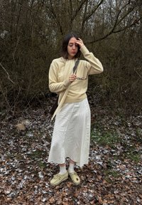 Jeune femme en pull beige et jupe blanche debout sur des feuilles givrées, tenant des lunettes et se touchant le front dans une zone boisée parsemée de feuilles.