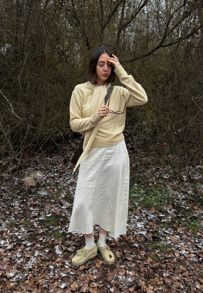 Joven mujer con suéter beige y falda blanca de pie sobre hojas heladas, sosteniendo gafas y tocándose la frente en un área boscosa cubierta de hojas.