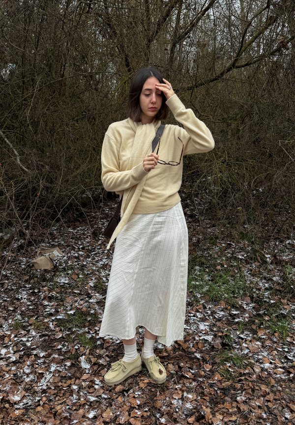 Mlada ženska v bež puloverju in beli krilu stoji na zmrznjenih listih, drži očala in se dotika čela v gozdnatem območju polnem listja.