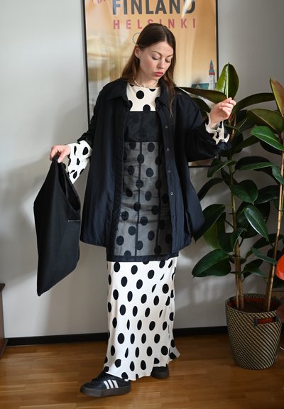 Chaqueta negra abotonada, superpuesta sobre un vestido negro con lunares y una falda blanca con lunares. Bolsa de mano negra, deportivas negras, plantas de interior al fondo.