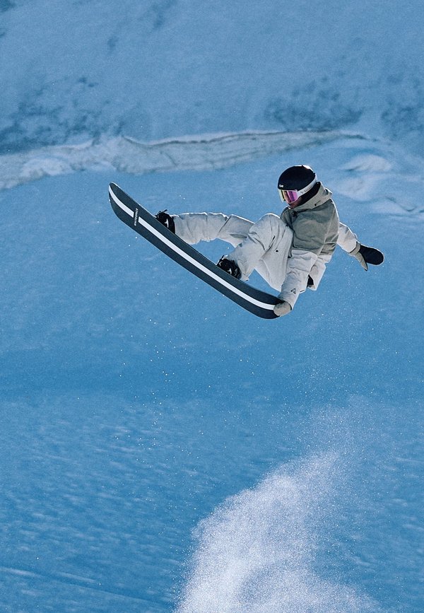 Un snowboardeur vêtu d'un équipement blanc et d'un casque réalise une figure en attrapant sa planche en plein saut au-dessus d'une pente enneigée, avec une pulvérisation de neige en dessous.