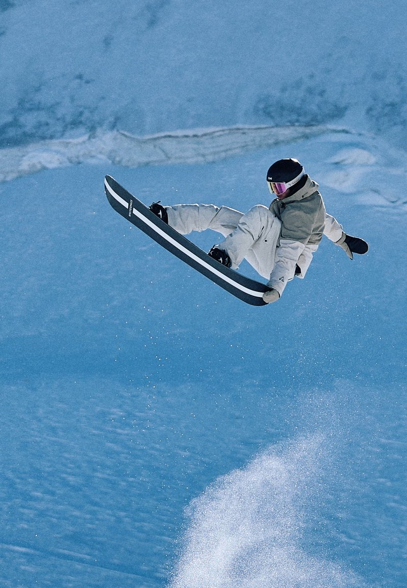 Σνόουμπορντερ με λευκό εξοπλισμό και κράνος εκτελεί κόλπο με άλμα και κράτημα στον αέρα πάνω από χιονισμένη πλαγιά με σπρέι χιονιού από κάτω.