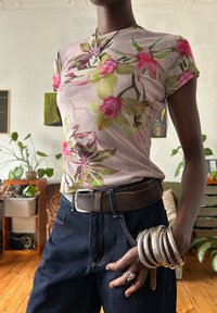 T-shirt com estampa floral em rosa claro, apresentando orquídeas verdes e rosas. Combinada com jeans escuros e um cinto de couro castanho. Múltiplos braceletes de prata.