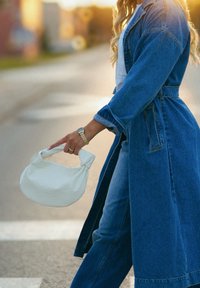 Bolsa texturizada branca segurada por uma pessoa com um casaco de ganga, apresentando uma alça redonda e um padrão subtil, em frente a um fundo de rua desfocado.