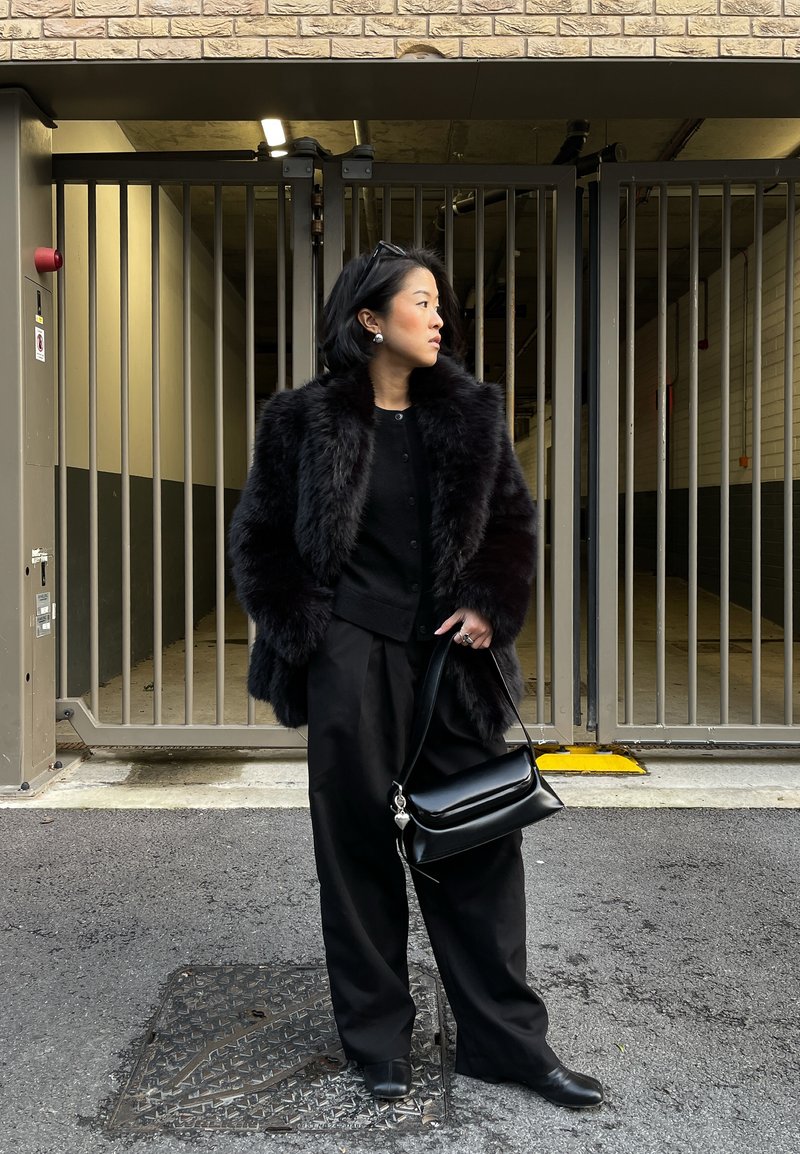 Femme en manteau de fourrure noir et pantalon large noir tenant un sac à main noir, debout sur l'asphalte devant une grille en métal surmontée d'un mur de briques.