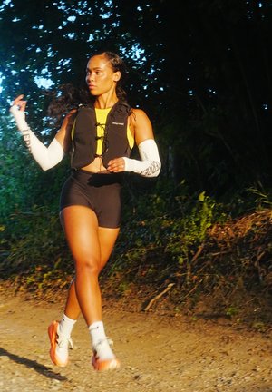 Chaleco ligero negro con cuerdas ajustables, camiseta amarilla, pantalones cortos negros y mangas de brazo blancas. Persona con zapatillas deportivas naranjas caminando por un sendero.
