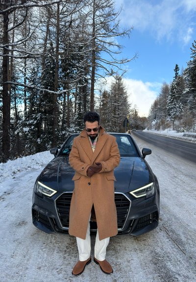 Hombre con abrigo marrón y guantes de pie frente a un coche negro en una carretera nevada, con árboles cubiertos de nieve y un cielo azul detrás de él.