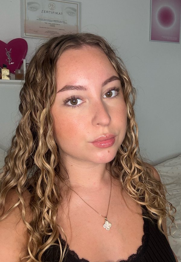 Jeune femme aux longs cheveux blonds bouclés portant un haut noir et un collier avec un pendentif en forme de trèfle, posant à l'intérieur avec des certificats sur le mur.