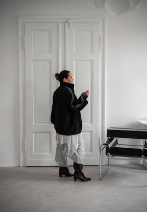 Mujer con cabello oscuro recogido en un moño, que lleva una chaqueta negra oversized, falda con capas y botas, está junto a puertas dobles blancas cerradas cerca de una silla negra.