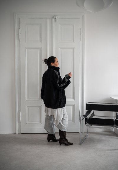 Mujer con cabello oscuro recogido en un moño, que lleva una chaqueta negra oversized, falda con capas y botas, está junto a puertas dobles blancas cerradas cerca de una silla negra.