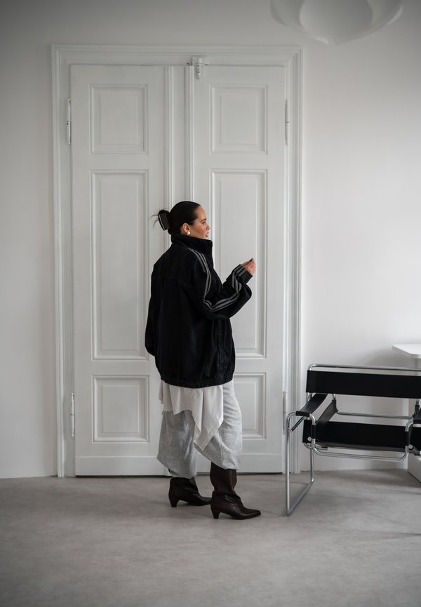 Femme aux cheveux foncés en chignon, portant une veste noire oversize, une jupe à couches et des bottes, se tient près de portes doubles blanches fermées, à côté d'une chaise noire.