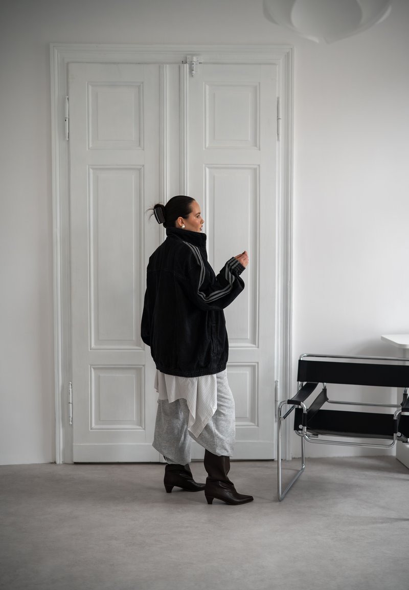 Žena s tmavými vlasy svázanými do drdolu, oblečená v oversized černé bundě, vrstvené sukni a botách, stojí u zavřených bílých dvoukřídlých dveří vedle černé židle.