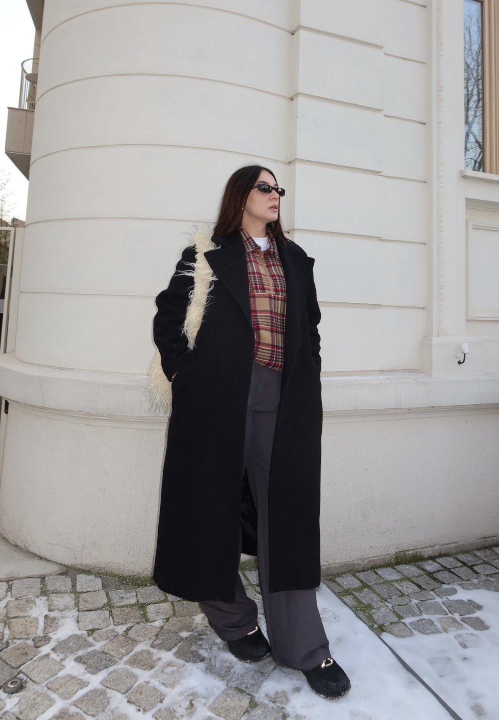 Femme portant un long manteau noir, une chemise à carreaux, un pantalon large gris, des lunettes de soleil noires et un sac à épaule blanc et duveteux, se tenant sur un pavé légèrement enneigé près d'un bâtiment blanc.