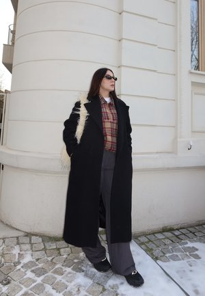 Mujer con abrigo largo negro, camisa de cuadros, pantalones anchos grises, gafas de sol negras y bolso blanco y esponjoso al hombro, de pie sobre adoquines con polvo de nieve cerca de un edificio blanco.