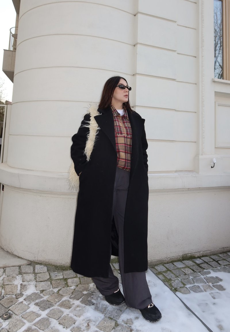 Femme portant un long manteau noir, une chemise à carreaux, un pantalon large gris, des lunettes de soleil noires et un sac à épaule blanc duveteux, debout sur des pavés saupoudrés de neige près d'un bâtiment blanc.
