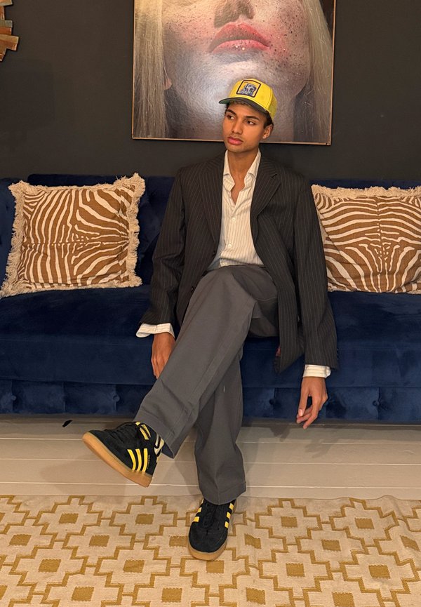 Giovane uomo con blazer a righe sottili, pantaloni grigi e cappellino giallo seduto a gambe incrociate su un divano blu con cuscini dalla stampa zebrata, sotto un grande ritratto in primo piano.