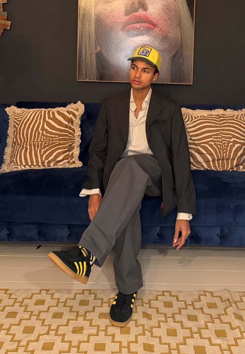 Tânăr cu blazer cu dungi subțiri, pantaloni gri și șapcă galbenă, stând în poziție cruce pe o canapea albastră cu perne cu imprimeu de zebre, sub un portret mare, realizat apropiat.