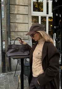 Brun tekstureret læderhåndtaske med to håndtag og en central lås, placeret på et bord; modellen har en beige kabelstrikket sweater og en mørk frakke på.