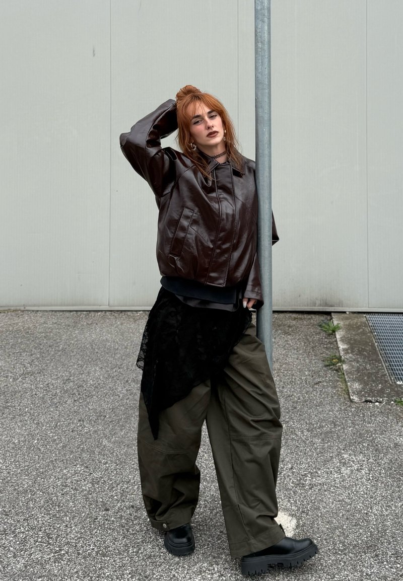 Braune Lederübersizejacke, schwarzer Schichtenstoff, lockere olivgrüne Hose und klobige schwarze Schuhe. Schlichter Hintergrund und städtische Umgebung.