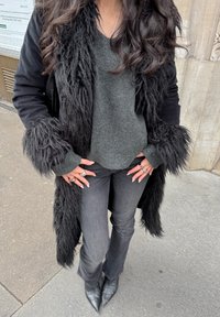 Žena v čiernom kabáte s kožušinovým lemom, tmavosivom svetri, čiernych džínsoch, špicatých čiernych čižmách a s viacerými striebornými prsteňmi na oboch rukách.