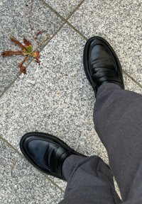 Zapatos de vestir de cuero negro con un acabado suave, que cuentan con una banda ancha sobre el empeine, combinados con pantalones grises, sobre una superficie de piedra.