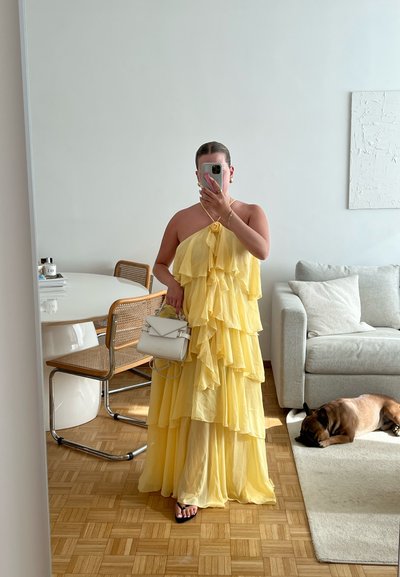 Vestido largo amarillos con volantes, de cuello halter, con capas transparentes y un adorno de flores, combinado con un bolso blanco y sandalias negras.