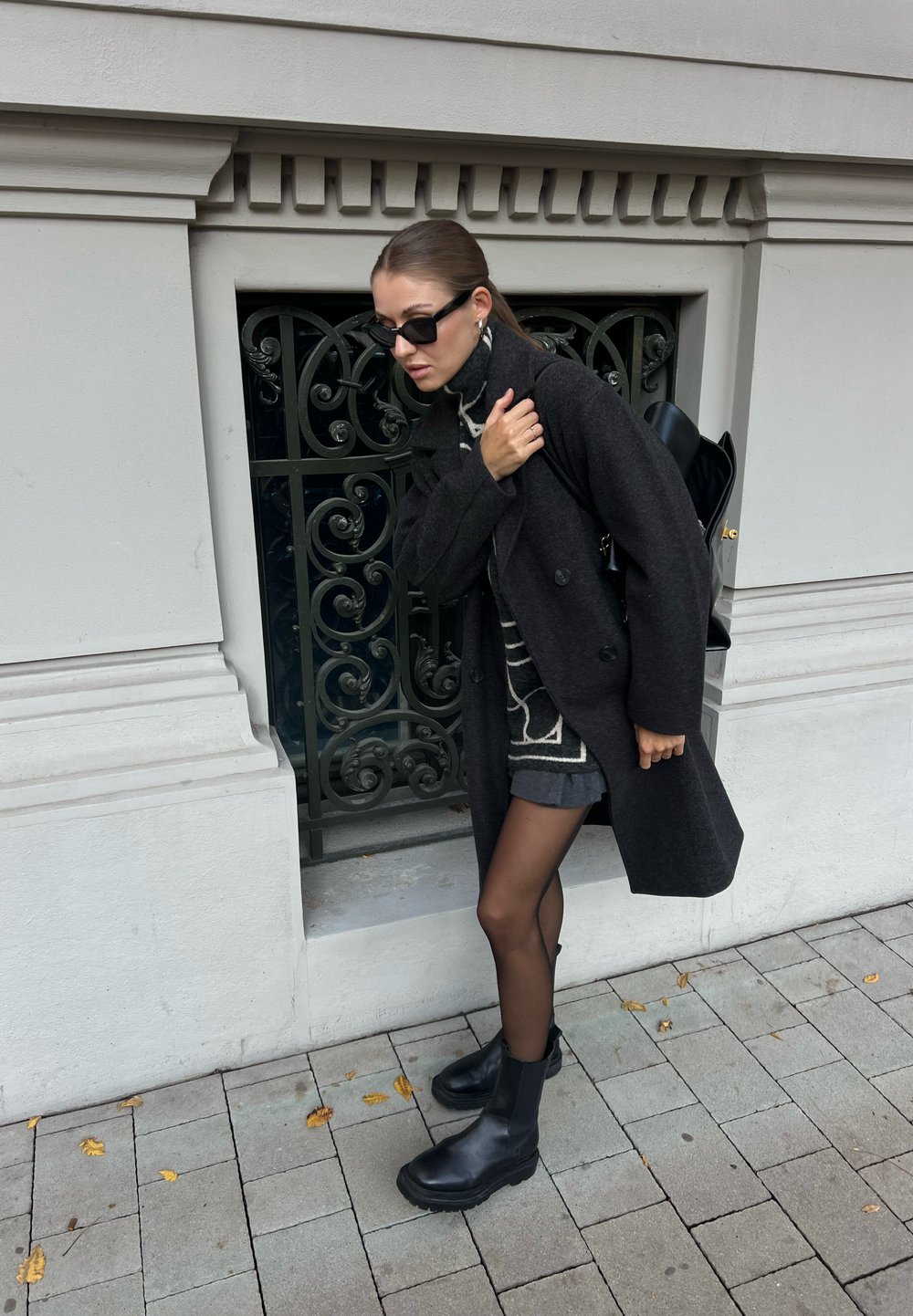 Cappotto di lana nero, maglione grigio a fantasia, pantaloni corti grigi, collant neri e stivaletti neri chunky. Piccolo zaino nero. Grata metallica ornata sullo sfondo.