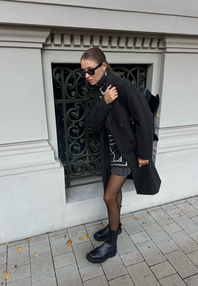 Abrigo de lana negro, suéter gris con patrón, pantalones cortos grises, medias negras y botines negros gruesos. Mochila negra pequeña. Reja de metal ornamentada detrás.