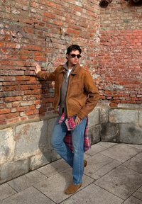 Jeune homme portant une veste en daim marron, un jean bleu et des lunettes de soleil, debout contre un mur en brique rouge usé en extérieur.