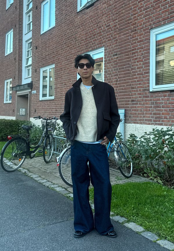 Veste noire portée sur un pull crème en maille, jean large foncé, et chaussures noires. Cadre extérieur avec des vélos et un mur en briques en arrière-plan.