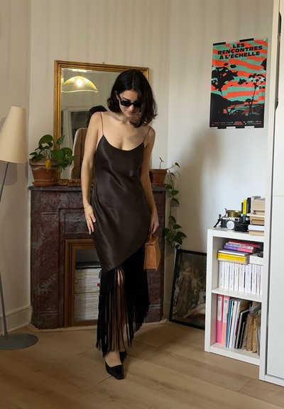 Vestido de satén marrón con tirantes finos, bajo asimétrico y detalles de flecos negros. Incluye un pequeño bolso de mano color camel y zapatos planos negros.