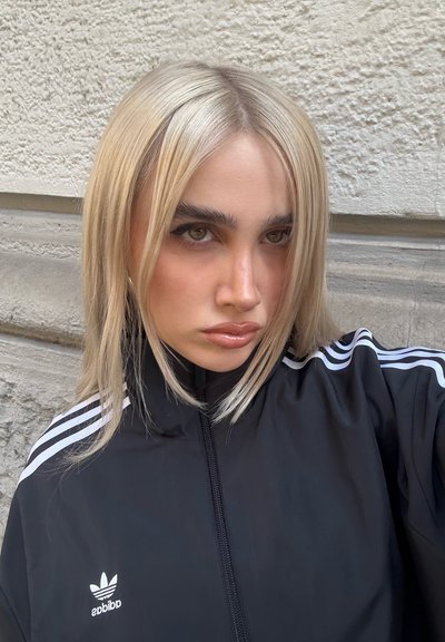 Chaqueta negra con cremallera y rayas blancas, con el logo de Adidas. Cabello rubio largo y recto enmarca la cara, con cejas definidas y labios brillantes.