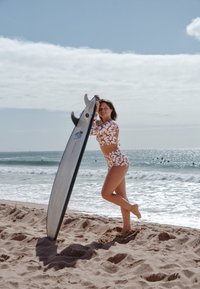 Daska za surfanje s oštrim vrhom, sivoj površini i crnim perajama, u kombinaciji s kupaćim kostimom bijele i narančaste boje s cvjetnim uzorkom, na pješčanoj plaži.